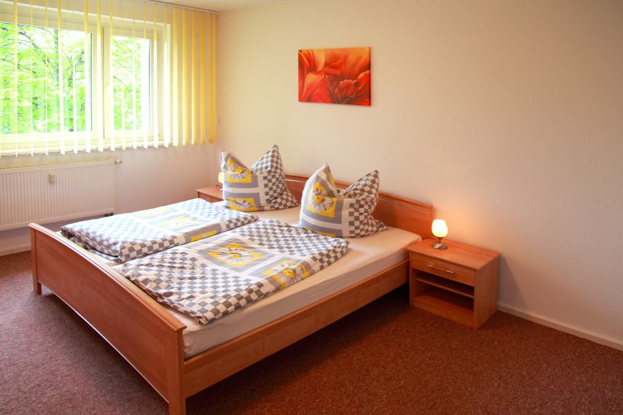 Schlafzimmer mit Doppelbett, Nachttischen, Teppichboden und Fenster mit Jalousien.