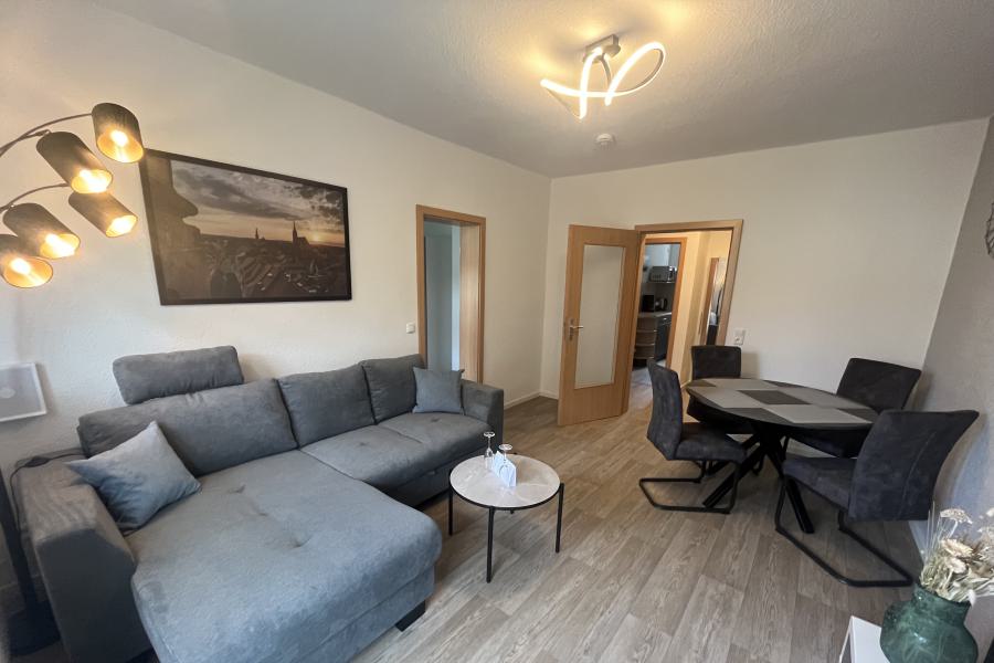 Wohzimmer mit Esstisch und Stühlen, Couch mit Bild darüber, Steh- und Deckenlampe und zwei geöffneten Türen.