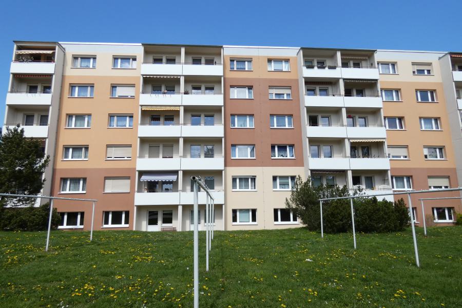 Mehrstöckiges Wohngebäude mit Balkonen, davor eine Grünfläche unter blauem Himmel.
