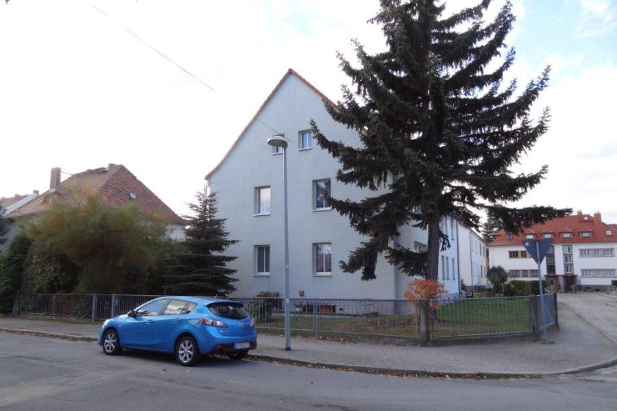 Kleines Mehrfamilienhaus an einer Straße stehend, ein Auto davor und ein hoher Nadelbaum.