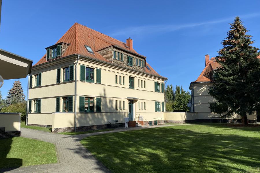 Zweistöckiges, historisches Wohngebäude mit grünen Fensterläden, einem Pflasterweg und Grünfläche davor.