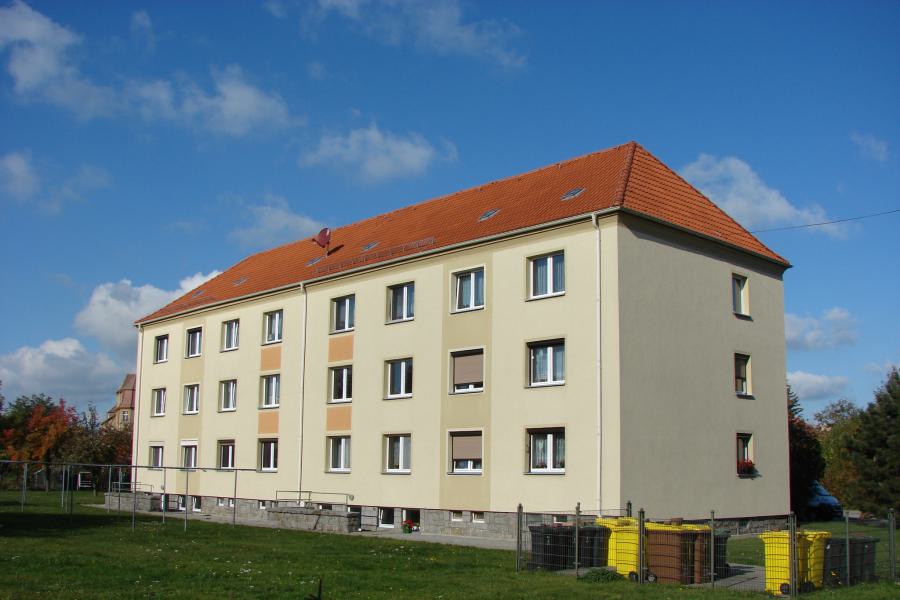 Dreistöckiges Mehrfamilienhaus mit Grünfläche davor und rechts ein Abfalltonnenbereich.