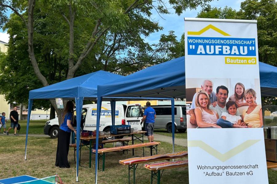 Eine Werbeplane für die Wohnungsgenossenschaft "Aufbau" Bautzen eG steht im Freien, daneben ein blaues Zelt, unter dem Tische und Bänke stehen. Im Hintergrund sind Bäume und Gebäude zu sehen. Die Werbeplane zeigt ein Bild einer lächelnden Familie und Kontaktdaten der Genossenschaft.