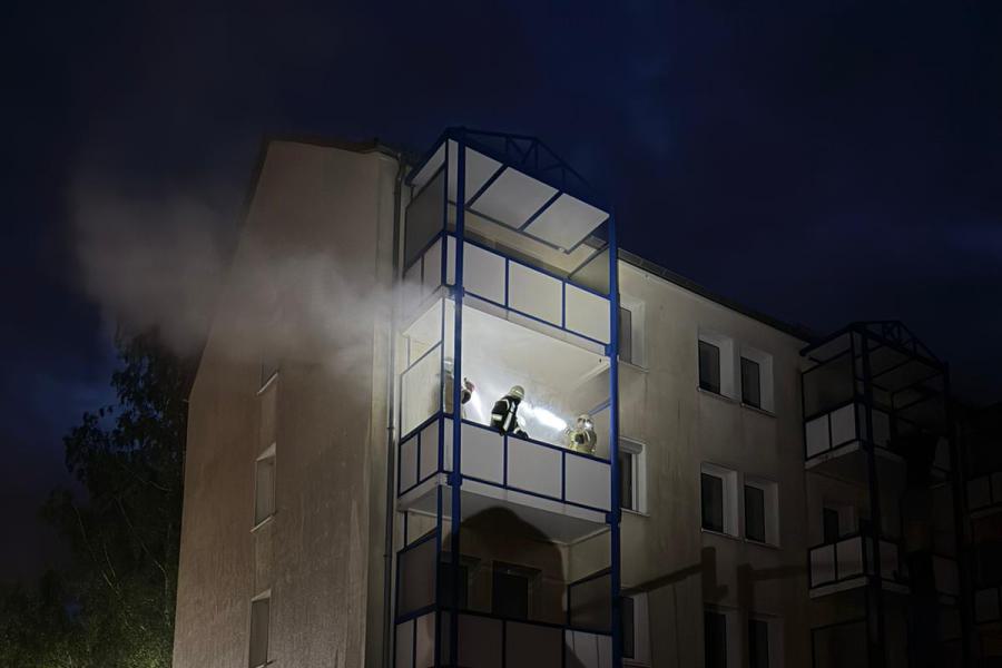 Nächtliche Aufnahme eines Mehrfamilienhauses während einer Feuerwehrübung. Aus einem oberen Balkon steigt künstlicher Rauch, während mehrere Feuerwehrleute das Gebäude mit Taschenlampen kontrollieren. Im Vordergrund steht eine Einsatzkraft in Schutzkleidung hinter einem Bauzaun.